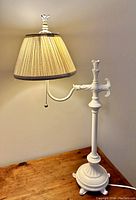 Full view of whitewashed wrought bedside lamp with beige pleated shade and pull chain on wooden surface against plain wall.