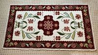 Full view of smaller rectangular handstitched crewel needlepoint rug showcasing the burgundy floral cross center, sage and dark green leaves, and floral border on a white background with looped fringe.