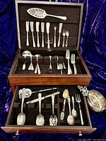 Full view of the open jeweler's wooden box with upper lid and lower drawer compartments displaying assorted flatware and the porringer to the side.