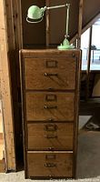 Front view of the vintage oak filing cabinet with four drawers and brass hardware, with the mint green adjustable desk lamp sitting on top.