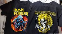 Photo showing front views of two black Iron Maiden T-shirts with colorful artwork featuring the mascot Eddie and text.