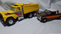 Side view of yellow Nylint dump truck and brown/orange ERTL pickup truck on white background.