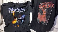 Two black t-shirts laying flat, one with Prince Purple Rain design on left, the other Michael Jackson Thriller design on right.