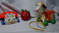 Photo showing 3 vintage Fisher Price toys: Chatter Telephone, Jingle Apple, Molly Moo cow pull toy on white cloth background.