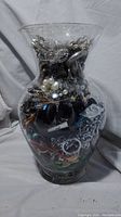 Front view of tall glass jar filled to the brim with assorted mixed jewelry including chains, bracelets, pendants, pearl-like beads and other jewelry pieces.