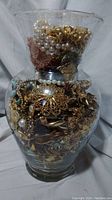 Full view of large glass jar densely packed with mixed costume jewelry pieces including faux pearls, gold-tone and silver-tone items.