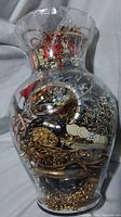 Large clear glass jar filled tightly with mixed jewelry of many types in gold and silver tones, showing collections of bracelets, rings, pendants, and more through the glass.