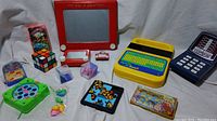 Photo shows complete assortment of small toys and brain games on white background, including Etch A Sketch, Rubik's Cube, and Speak & Read toy.