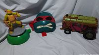 Three items on a white cloth background: TMNT Michelangelo figure, red bandana shell mask, and remote control shell vehicle with camouflage colors.