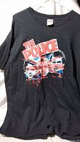 Front view of black T-shirt showing The Police band members' faces and logo in red, white and blue splatter paint style.