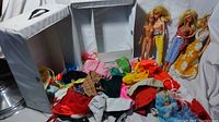 Overall view of Barbie doll trunk open showing 4 Barbie dolls standing inside and many pieces of clothing scattered in and beside trunk.
