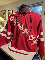 Front view of red Canada hockey jersey with 'CANADA' lettering and captain's 'C' patch, lace-up collar visible.
