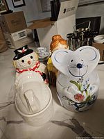 Photo showing four ceramic cookie jars including a snowman, turkey, gramma, and mouse on a kitchen counter with some surrounding boxes and kitchen items.