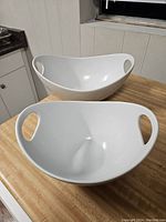 Two large white ceramic serving bowls on a wooden counter, showing oval shapes with integrated handles on both sides.