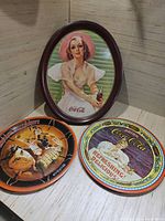 Three trays in one photo: large oval Coca-Cola tray with woman image, round Coca-Cola tray with polar bear, and round Coca-Cola tray with retro couple.