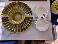Four-piece ceramic serving tray set displayed on a countertop, showing the three nested avocado green trays on the left and a white double shell-shaped tray with central metal handle on the right.