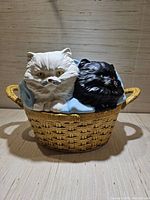 Front view showing ceramic container designed to look like a wicker basket with ceramic white and black cats inside on blue cloth.