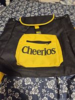 Front view of black backpack with yellow front zippered pocket labeled Cheerios and top flap closure with yellow trim and buckle.