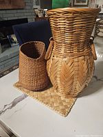Two wicker baskets shown together, one smaller smooth weave and one taller decorative weave with handles, displayed on a woven placemat on a table.