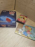 Three game items including a boxed English Murder Party Game, a small colorful card game box, and a vintage children and lamb puzzle.