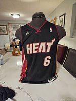 Front view of black Miami Heat LeBron James basketball jersey, size small, on a mannequin. Shows Adidas and NBA logos, 'HEAT' text, and number 6.