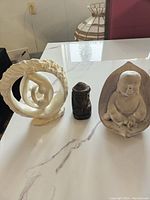 Full lot: three carvings including the white abstract stone carving, small dark carved head, and cement Buddha statue arranged on a white marble table.