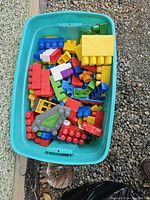 Turquoise tub filled with multicolored Duplo blocks in various shapes and sizes, placed on a gravel surface.