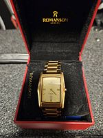 Front view of Romanson gold plated watch in its box showing rectangular gold-tone dial with Roman numeral markers and date window.