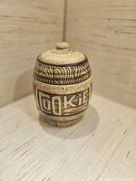 Photo of a midsize ceramic cookie jar with wood-grain texture and 'COOKIE' engraving on the front, beige and brown coloration, displayed on a light wood surface.