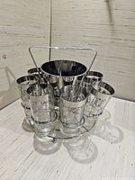Full set showing six silver plated glasses arranged around a silver plated ice bucket, all held on a metal stand against a light wooden surface and background.