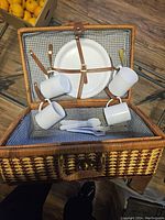 Open wicker picnic suitcase showing four white plates, four white cups, and cutlery inside secured with leather straps on gingham fabric lining.