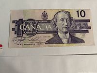 Front view of worn 1989 Canadian $10 bill with crease and visible portrait of Sir John A. Macdonald, and Parliament buildings in background.