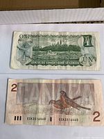 One $1 and one $2 Canadian paper bill, reverse sides showing denomination and design features.
