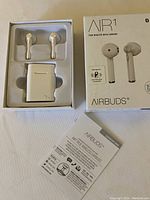 Open box with white wireless earbuds, charging case inside molded packaging, box cover, and user manual.