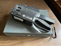 Black Clary 324-010 price labeller resting on gray metal cash box, showing labeller handle and part of tape feed.