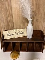 Wooden letter holder with front compartments, white ceramic vase with feather plume, 'Laugh Out Loud' tin sign, and amber glass paperweight arranged on wooden floor background.