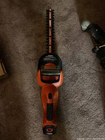 Top view of hedge trimmer showing long cutting blade with dual-sided teeth and orange handle with black grip on carpet.