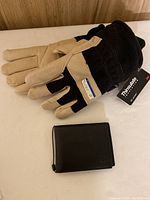Pair of men’s black and beige Thinsulate gloves with visible tag and new black leather wallet closed on a white surface.