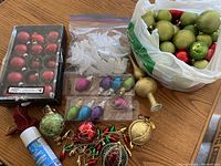 Top-down photo of assorted Christmas ornaments including small red glass balls in box, white leaf-shaped plastic ornaments, colorful small ribbed glass teardrop ornaments, and a white bag filled with assorted round ornaments in green, red, and gold.