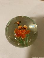 Close-up of round glass paperweight with an orange flower and bee inside, showing clear glass with small air bubbles.