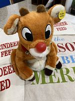 Close-up of vintage Rudolph plush reindeer with red nose and brown antlers. Plush has small battery pack and tag 'PUSH HERE'.