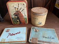 Four vintage tins displayed on wooden surface: Player's Navy Cut Cigarettes tin, Black Cat Virginia Cigarettes tin, Pictman round tobacco can, and bagpiper rectangular tin.