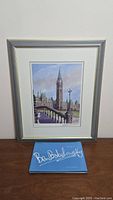 Framed signed and numbered print of Parliament Hill with art book beneath it on a wooden surface.