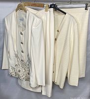 Three vintage women's formal clothing sets hung on hangers, showing the full view of each jacket and skirt. The Vintage Lucia set has embroidered floral design with rhinestone accents and decorative buttons. The other two sets are cream-colored with detailed buttons and textured design.
