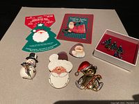 Photo of all seven Christmas-themed pins and brooches arranged on flat surface showing individual details and original cards for two pins.