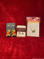 Photo shows three jewelry items against a red fabric background: colorful Pooh earrings on black cards, black and white Mickey Mouse stud earrings in a cream jewelry box, and a Lion King necklace with a Simba pendant in a clear package with branded backing.