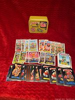 Top view of the Garbage Pail Kids cards fanned out in front of the yellow collectible tin box on red fabric.