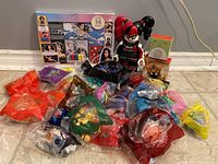 Lot of McDonald's toys with Harley Quinn clock figure and Wonder Woman coloring set, assorted toys in colored plastic bags arranged on floor against wall.