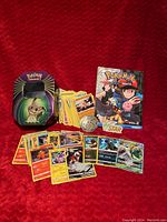 Photo of lot items arranged on red textured fabric background showing the Pokémon tin, various trading cards, metal coin, and collection book.