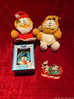 Photo showing 2 Garfield Christmas plush toys, Frosty the Snowman ornament in box, and a McDonalds Disney snow globe on red sled base.